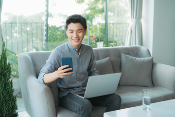 Wall Mural - Smiling young man typing on mobile phone while sitting on a sofa at home with laptop computer