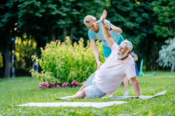 Happy gray-haired pensioner father and Young woman physiotherapist help him doing side plank on one hand exercise on mat at summer park, outdoor. Active lifestyle and healthcare in any age concept..