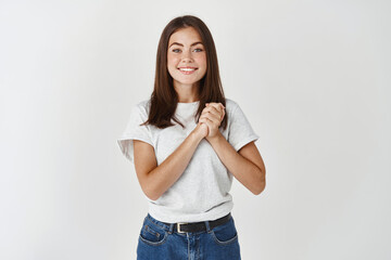 Grateful young woman holding hands together and looking at camera hopeful, saying thank you, standing delighted against white background