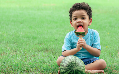 Portrait of little african curly hair cute boy playing, openning large mouth to eat piece of watermelon with happiness while sitting on the green field in park. Fruits, Food and Education Concept.