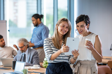 Wall Mural - Young colleagues coworkers working together in company meeting room using digital tablet. Smiling businesswomen using digital tablet. Female business professionals are working on project. 