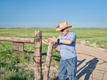 Senior Man In A Cowboy Hat Is Opening Or Closing Cattle Barbed Wire Gate On A Ranch Road, Summer Scenery With Green Prairie In Colorado