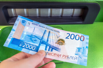 Man's hand holds new Russian banknote of two thousand rubles against background of green cash machine