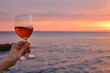 © Iuliia Pilipeichenko - A glass of red wine in the hands of a girl in the rays of the sunset on the seashore. Alcoholic beverage tasting. Beautiful sunset with wine on the beach. Romantic evening aperitif. Glass closeup