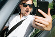 © Sergio - Portrait of a surprised young man with an open mouth filming an event on a mobile phone while driving a car. Selective focus on the shocked face of the driver.