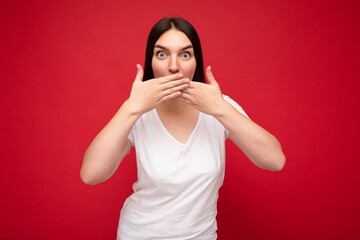 Portrait of young pretty nice brunette female person with sincere emotions wearing casual white t-shirt for mockup isolated over red background with copy space and covering mouth with hands