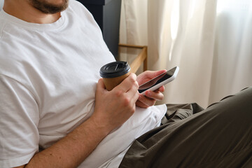Wall Mural - a man holds a phone and a cup of coffee in her hands. a man sits on the sofa and having fun with a g