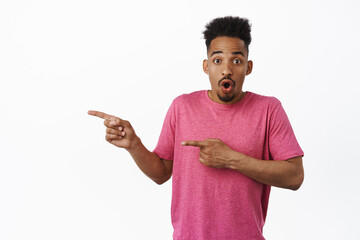 Portrait of surprised african american man gasp, say wow and stare amazed, pointing fingers left at sale discount logo, showing advertisement, standing in pink t-shirt over white background