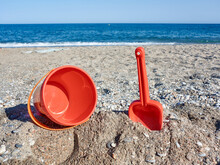 Red Plastic Shovel And Bucket For Children In The Sand On The Beach In Front Of The Sea, Iconic Image Of Summer And Vacation Period     