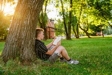A Young Man Is Sitting Under A Tree And Reading A Book. Green Summer Park At Sunset