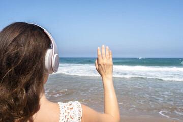 Happy woman in headphones listen music waving hand to say hello and welcoming sea standing on the beach. Summer vacation. Back view