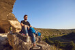 © Studio Romantic - Young hiker sitting and having rest on natural rocks and enjoying green valley landscape with his group at background during summer vacations on sunny clear day. Hiking and traveling concept
