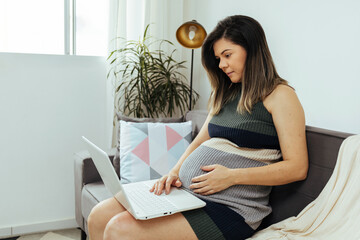 Pregnant woman sitting on sofa using laptop. Concept of pregnant woman working from home