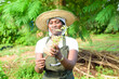 African female gardener, florist or horticulturist wearing an apron and a hat, holding a bag of plant as she works in a green and colorful flowers garden
