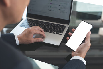 Canvas Print - Mockup image, business man hand holding black black mobile smart phone  working on laptop computer on table at office