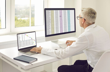 Senior accountant man using laptop and desktop computer while working in modern office. Mature white male company employee and team project manager sitting at desk, looking at screen 