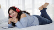 © Bangkok Click Studio - Asian young happy pretty friendly fat oversized woman lay down smiling relaxing on white sheet bed listening to music from big red earphones use playlist from black tablet on hand at home in bedroom