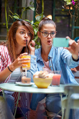 Wall Mural - Two young and beautiful female friends making funny grimaces while taking a selfie in the bar. Leisure, bar, friendship, outdoor