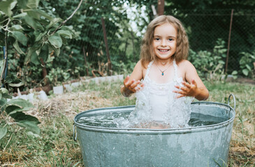 little cute girl in a pink swimsuit flops, having fun and splashing in a basin in the summer in the backyard, the concept of a happy childhood and summer vacation