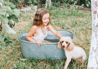 little cute girl in a pink swimsuit flops, having fun and splashing in a basin in the summer in the backyard with poodle dog, the concept of a happy childhood and summer vacation