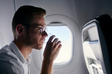 Wall Mural - Portrait of passenger during flight. Young man drinking water from plastic cup.