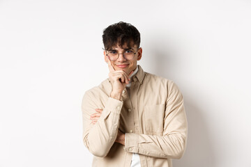 Cunning young man watching something interesting happen, lean face on arm and smiling devious, looking at camera intrigued, standing on white background