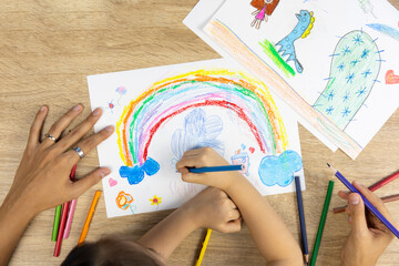 Top view shot of mother and daughter are drawing and coloring beautiful rainbow picture on paper together shows concept of art playing which enhance creativity, learning and enjoyment for child.