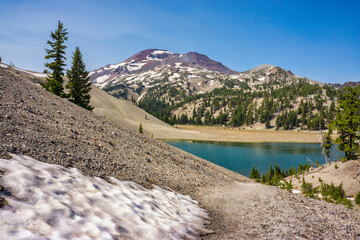 Wall Mural - patch of melting snow and a clear blue alpine lake surrounded by pumice covered moraine, fir trees, 