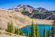 Clear blue alpine lake surrounded by pumice covered moraine, fir trees, and volcanic mountain peaks.