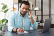 © kucherav - Distance work concept. Young indian man support worker or freelancer uses laptop and headset to communicate with the client, makes notes in a notebook, online learning