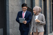 © NVB Stocker - Young business muslim woman and business man colleague walking and discussing outside the office. Woman is holding a coffee and businessman is holding a digital tablet outdoor.