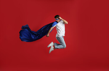 Wall Mural - Man in superhero cape and mask jumping on red background