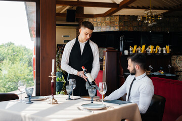 A young waiter in a stylish apron demonstrates and offers a fine wine to a client in a restaurant. Customer service.