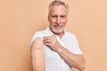 Old Bearded Man Got Vaccinated Against Coronavirus Shows Arms With Adhesive Plaster Cares About Healthy During Pandemic Poses Against Brown Background. After Vaccination And Immunization Concept.