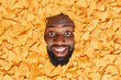 Cheerful dark skinned man with thick beard buried in delicious mexican nachos chips enjoys eating tasty salty spicy snack smiles broadly. Positive guy surrounded by triangular crunchy crisps