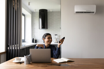 Poster - Happy female Indian student or employee working from home at workplace with laptop, using remote control for regulating air conditioner, cooling hot air, relaxing under cold breeze. Comfort concept