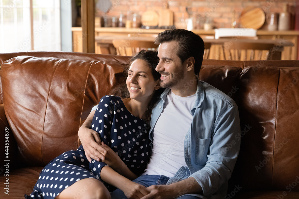 Happy young family couple relaxing on leather couch at home or in stylish hotel apartment. Dating man and woman in love hugging on sofa, talking and smiling, looking away, thinking over good future - obrazy, fototapety, plakaty 