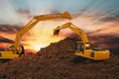 Two excavators are digging the soil in the construction site on the sunset sky background,With bucket lift up