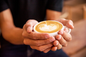 Wall Mural - Close up hands of man sitting office desk holding sweet coffee cup relax and enjoy with happy time. Hot coffee mug in hand. Man holding coffee cup relaxing after work at office warm taste in cafe