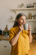 © shurkin_son - Smiling middle-aged woman drinking tea in home kitchen. Cheerful elderly female enjoying morning coffee. Happy housewife looking away with gladness. Modern kitchen interior on blurred background