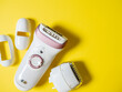Female electric epilator in white on a yellow background. There are additional attachments nearby. Top view, flat lay