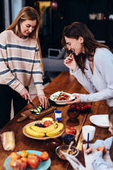 Two young women are preparing lunch, one is cutting vegetables for a salad, the other is tasting and the girl next to her is doing her homework.