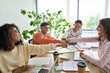 © insta_photos - Young happy laughing creative startup team four multiethnic coworkers diverse students work together give fist bump celebrate successful project in office classroom at desk. Teamwork success concept.