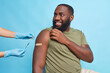 © wayhome.studio  - Cheerful bearded adult Afro American man got covid 19 vaccine looks with happy expression at nurse shows shoulder with adhesive tap poses against blue background. Coronavirus vaccination concept