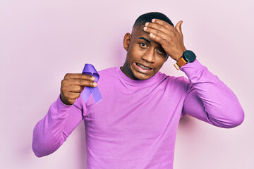 Young black man holding purple ribbon awareness stressed and frustrated with hand on head, surprised and angry face