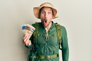 Sticker - Handsome man with beard wearing explorer hat holding canadian dollars scared and amazed with open mouth for surprise, disbelief face