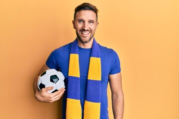 Wall Mural - Handsome man with beard football hooligan cheering game holding ball looking positive and happy standing and smiling with a confident smile showing teeth