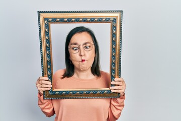 Wall Mural - Middle age hispanic woman holding empty frame making fish face with mouth and squinting eyes, crazy and comical.