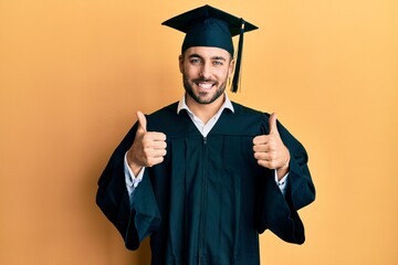 Sticker - Young hispanic man wearing graduation cap and ceremony robe success sign doing positive gesture with hand, thumbs up smiling and happy. cheerful expression and winner gesture.