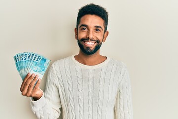 Canvas Print - Handsome hispanic man with beard holding 100 brazilian real banknotes looking positive and happy standing and smiling with a confident smile showing teeth